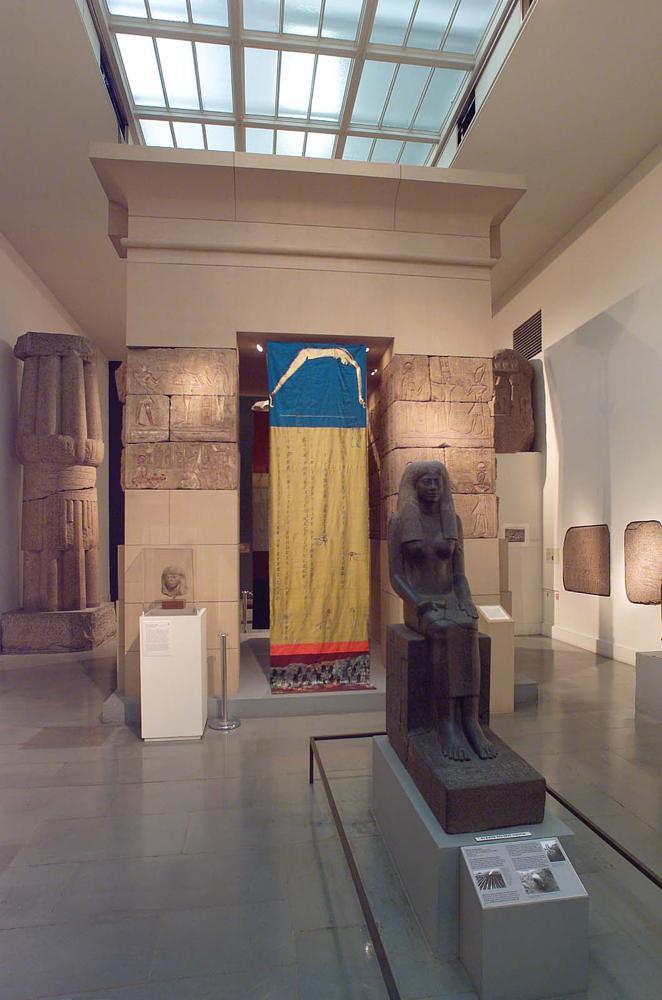 A printed and stitched textile hangs in a portal space in an Egyptian gallery. A sculpture of a seated Egyptian woman is in the foreground.
