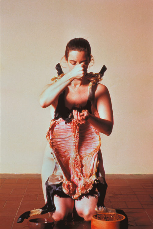 A woman with her head slightly bowed and hair pulled back. She wears an animal carcass, its legs rest on her shoulders. Her right hand is raised and she pours salt from it into her left hand.