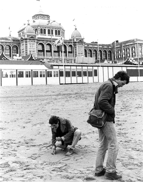 Two male figures seen on a sandy beach, one is bent over the sand and is picking it up, the other is turned to the right and his slightly hunched with his hands in his pockets. Behind them is an ornate building.