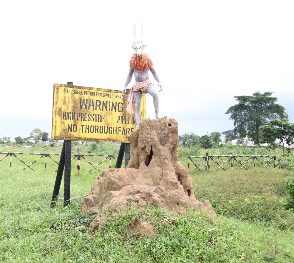 A man in a mask stands atop a termite mound in front of a sign reading Warning High Pressure Pipeline No Trespassing