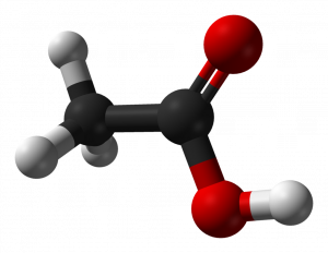 Ethanoic acid as a ball and stick image. The molecule includes 2 connected black spheres representing carbons. On the first are 3 small white spheres representing hydrogen atoms. The other carbon is attached to two red spheres, one by a double bond and the other by a single bond, and with a white hydrogen sphere attached to it.