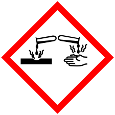 Pictogram is a white diamond outlined in red, with an image showing a test tube to the left pouring liquid onto a solid surface, and another test tube to the right pouring a substance onto a hand. Squiggly lines emanating from the surface and the hand indicate damage or injury.