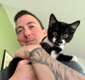 Emerson, who has short brown hair and a tattoo of a tree on their forearm, is holding a black and white kitten named Ranger. The background is green.