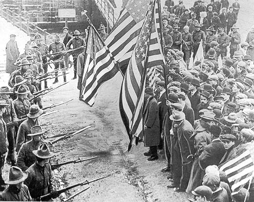 Soldiers point rifles with bayonets at a crowd of strikers holding American flags