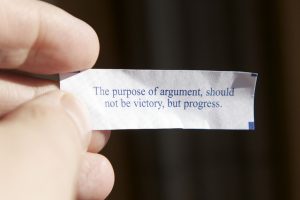 a picture of a hand holding a fortune from a fortune cookie; the fortune reads "The purpose of argument should not be victory, but progress."