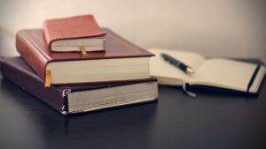 a stack of books next to an open book and pen on a desktop