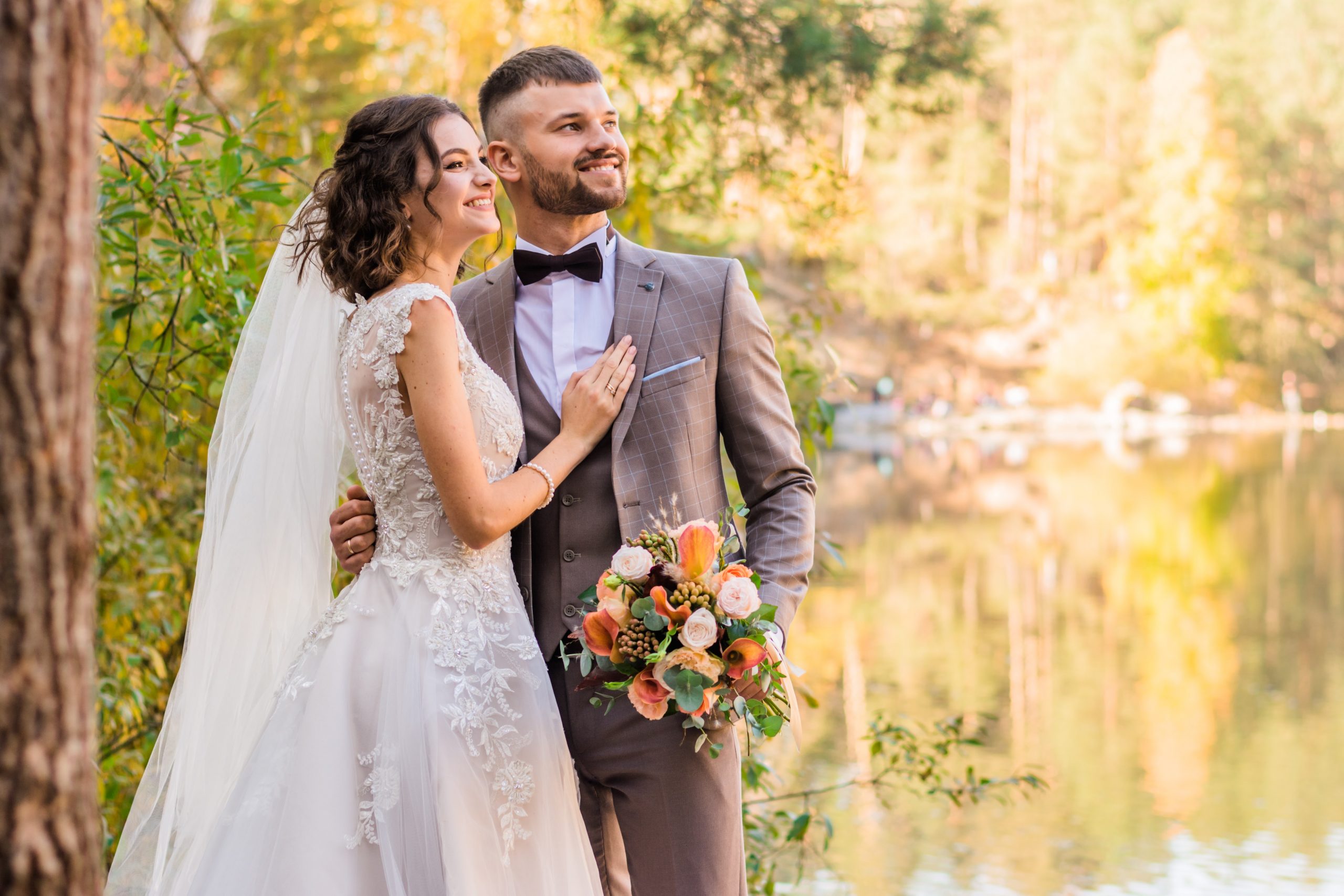 Photograph of a woman in a wedding dress and a man in a tuxedo standing near a body of water.