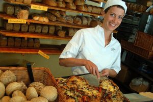 Woman working in a bakery, smiling as she puts baked breads on display.