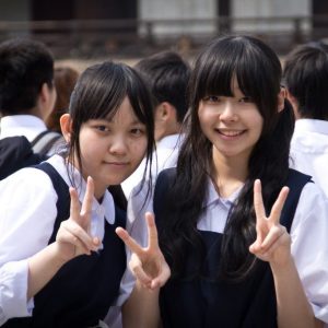 Two Japanese school girls smile and flash a typical pīsu or &quot;peace sign&quot; as the pose for a photo.