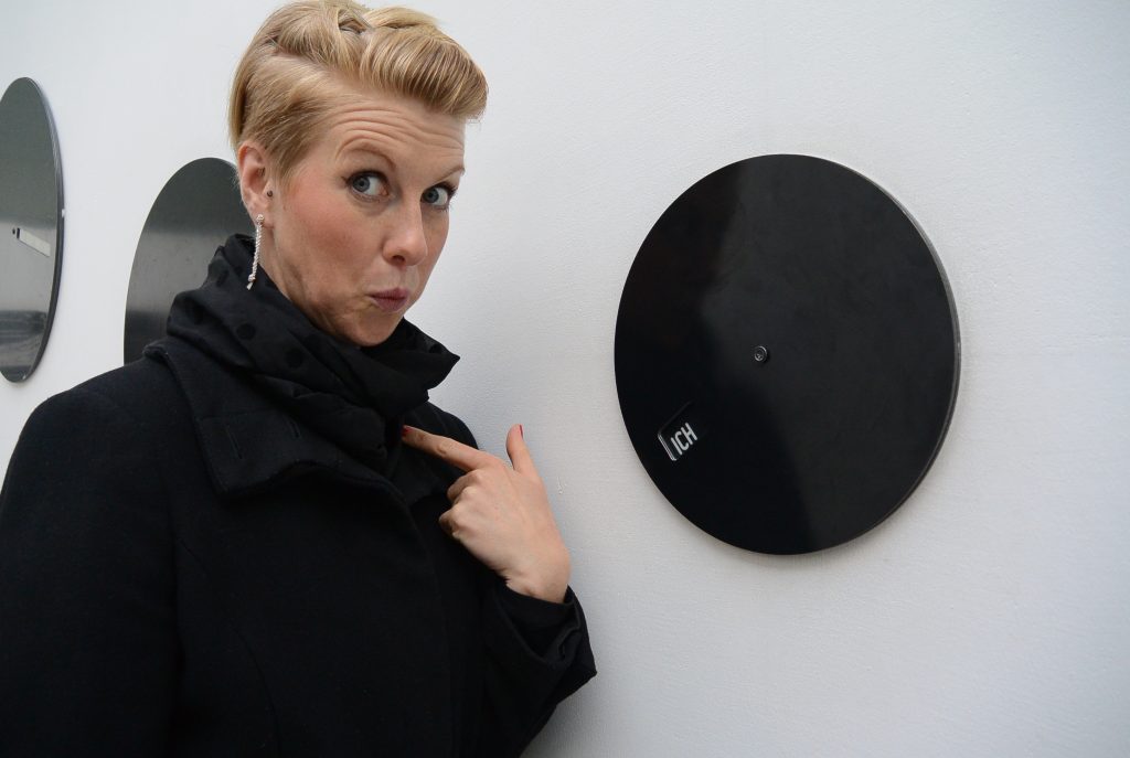 A feminine presenting person pointing to themself next to a record disk mounted on a wall.