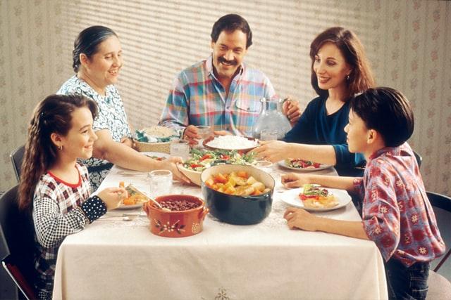 A grandma, mom, dad, son, and daughter all gathered around a family dinner table dishing up food, eating, and smiling.