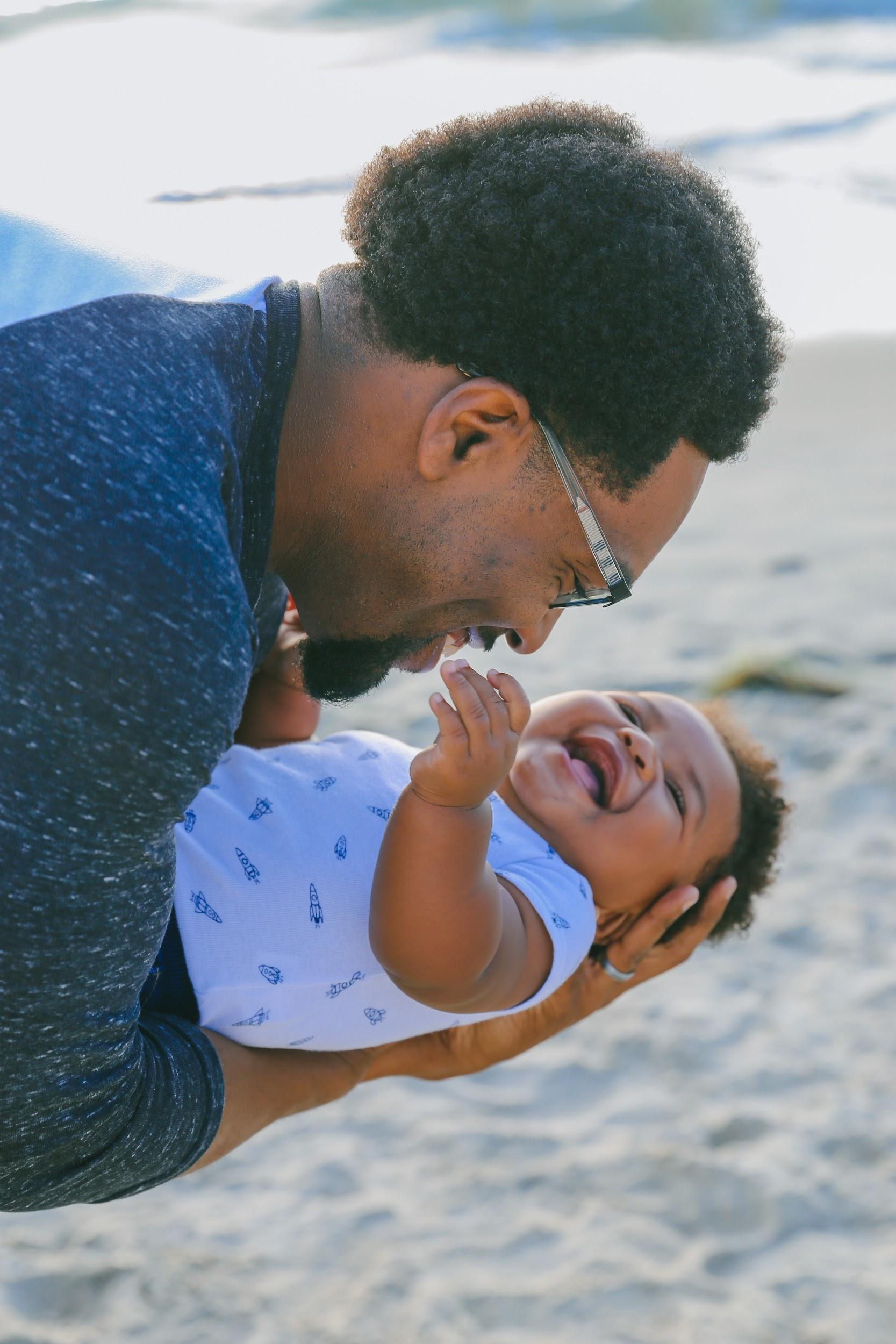 A Black father with facial hair and glasses holds his baby playfully on the beach. The father is smiling warmly, and the baby is laughing.