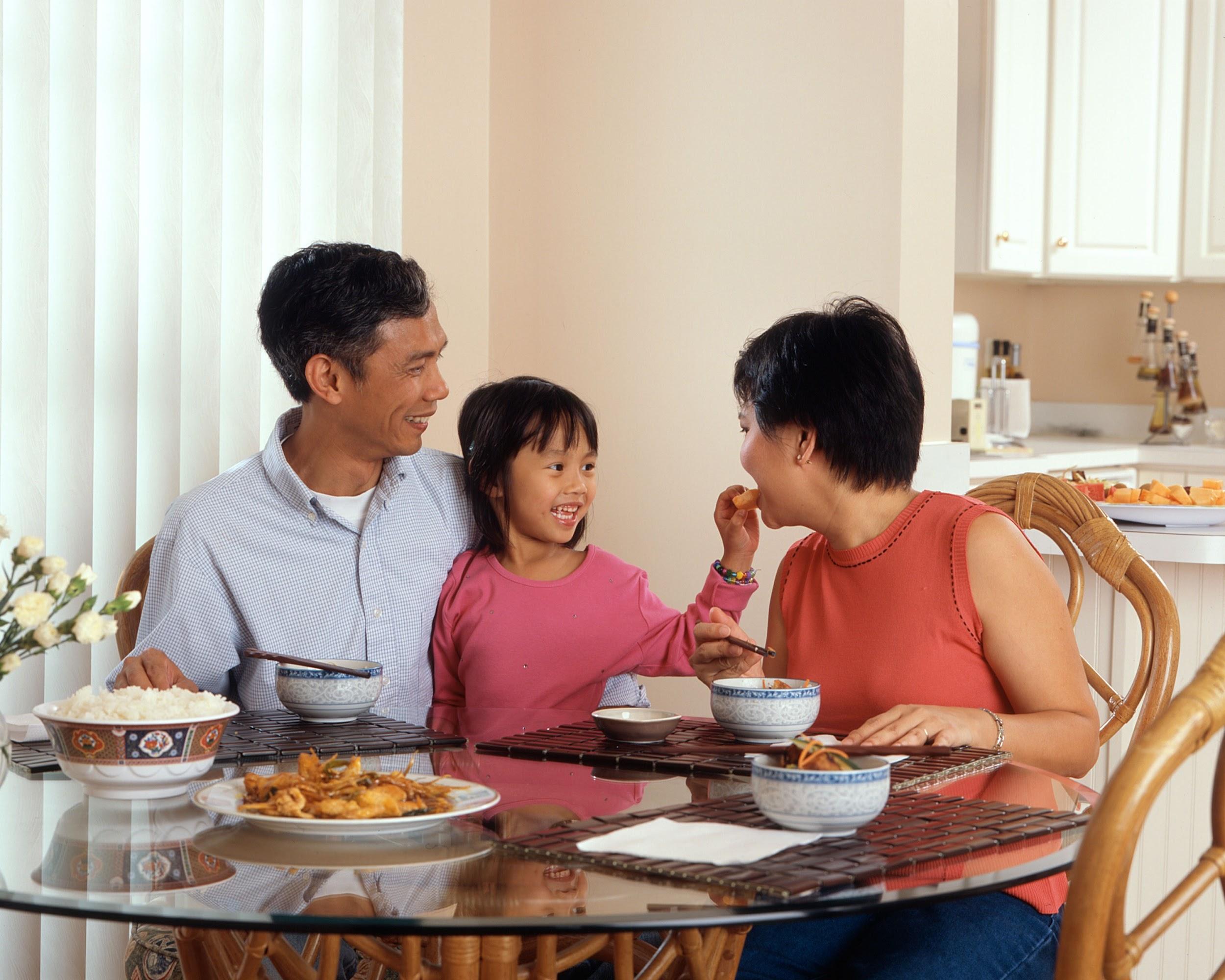 A family of three (mom, dad and daughter) sitting at a glass round table, enjoying a meal of rice and chicken. The young child is feeding the mom a bite of food.