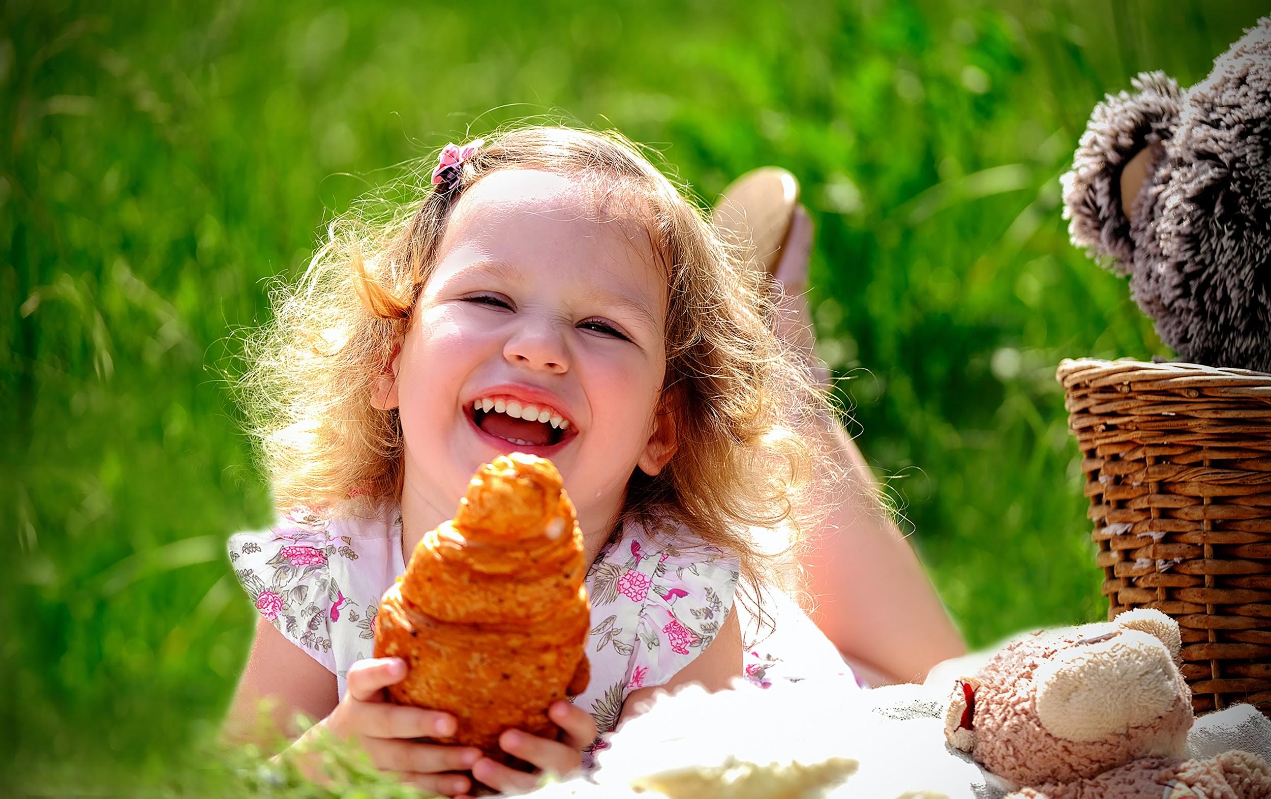 A little girl laying in a field on her tummy. She is looking at the camera smiling and laughing, and holding a croissant.