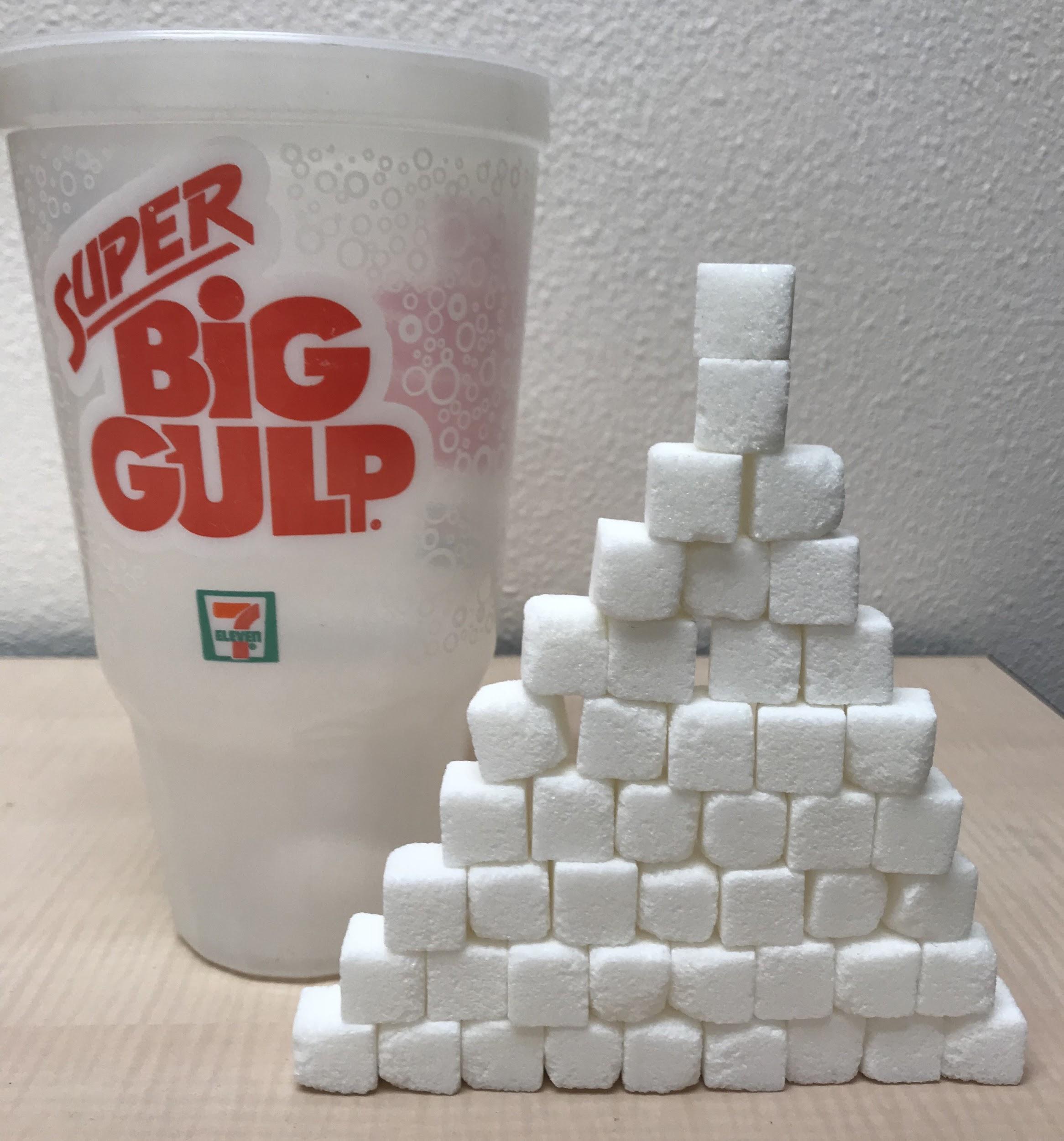 Forty six sugar cubes stacked next to a big gulp to illustrate the 46 teaspoons of sugar that the soda contains.