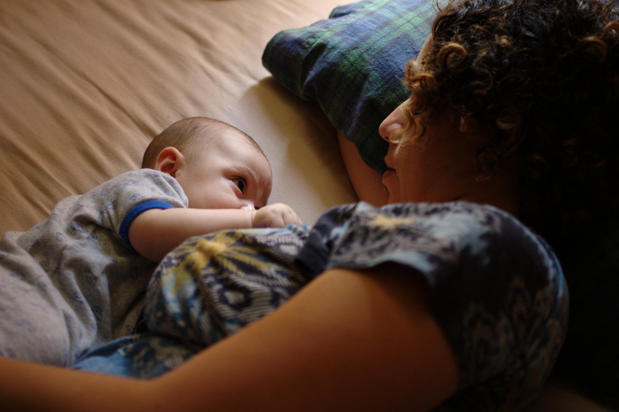 A mother is shown lying comfortably on her side, head propped up by a pillow, with her baby lying next to her and breastfeeding. Mother and baby are making eye contact.