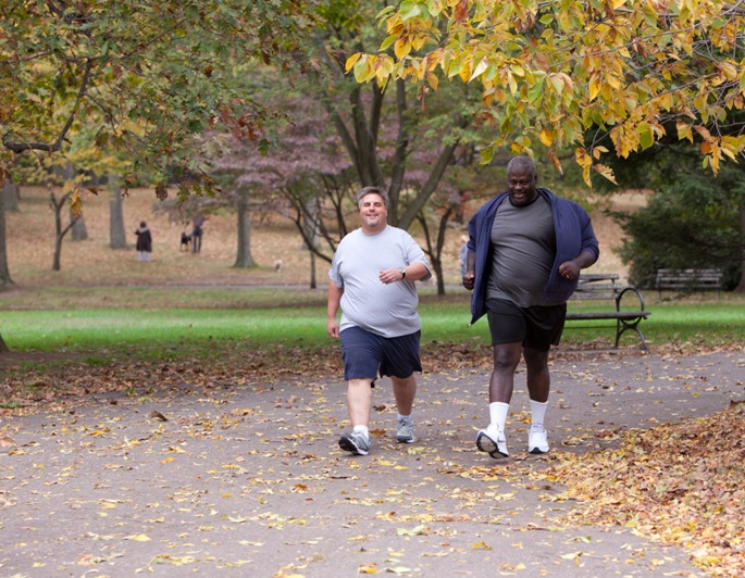 Two men walking down a path in a park with fall leaves on the trees and ground.