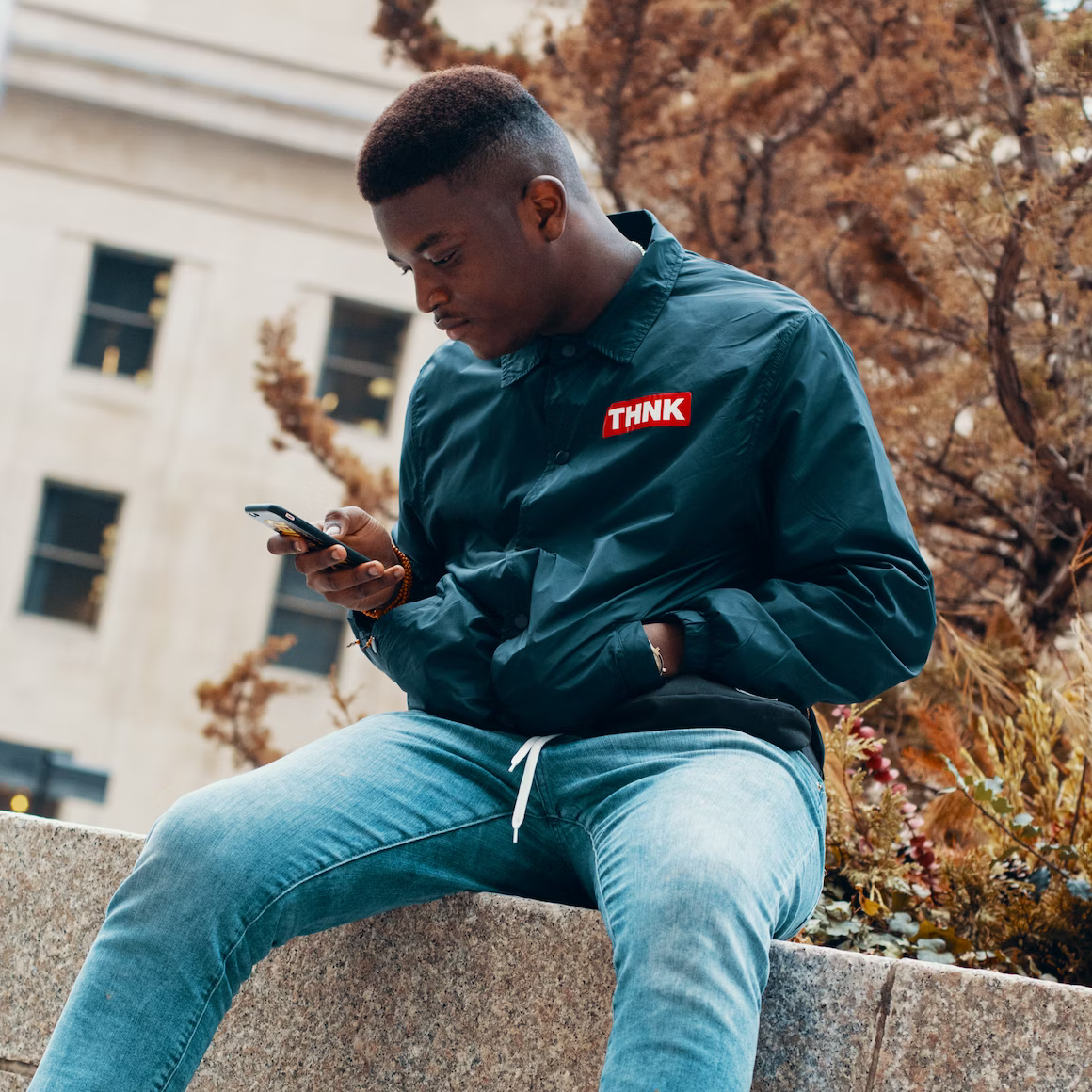 A black man sitting on a stone wall looking at his phone.