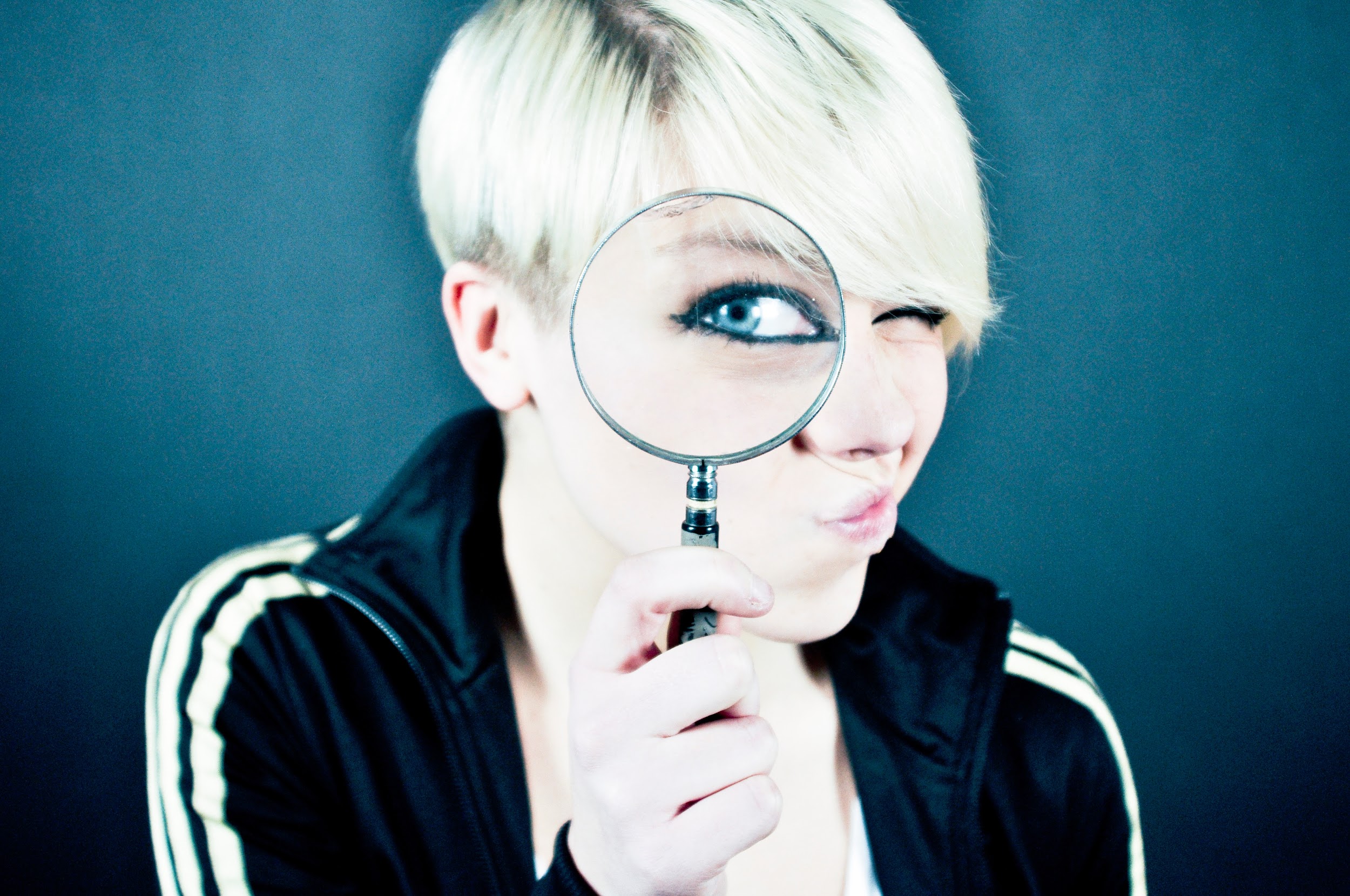 A young woman with short dyed blonde hair and wearing a black and white track jacket peers through a magnifying glass. Through the glass, we can see her blue eye, its edges heavily darkened with eye liner.