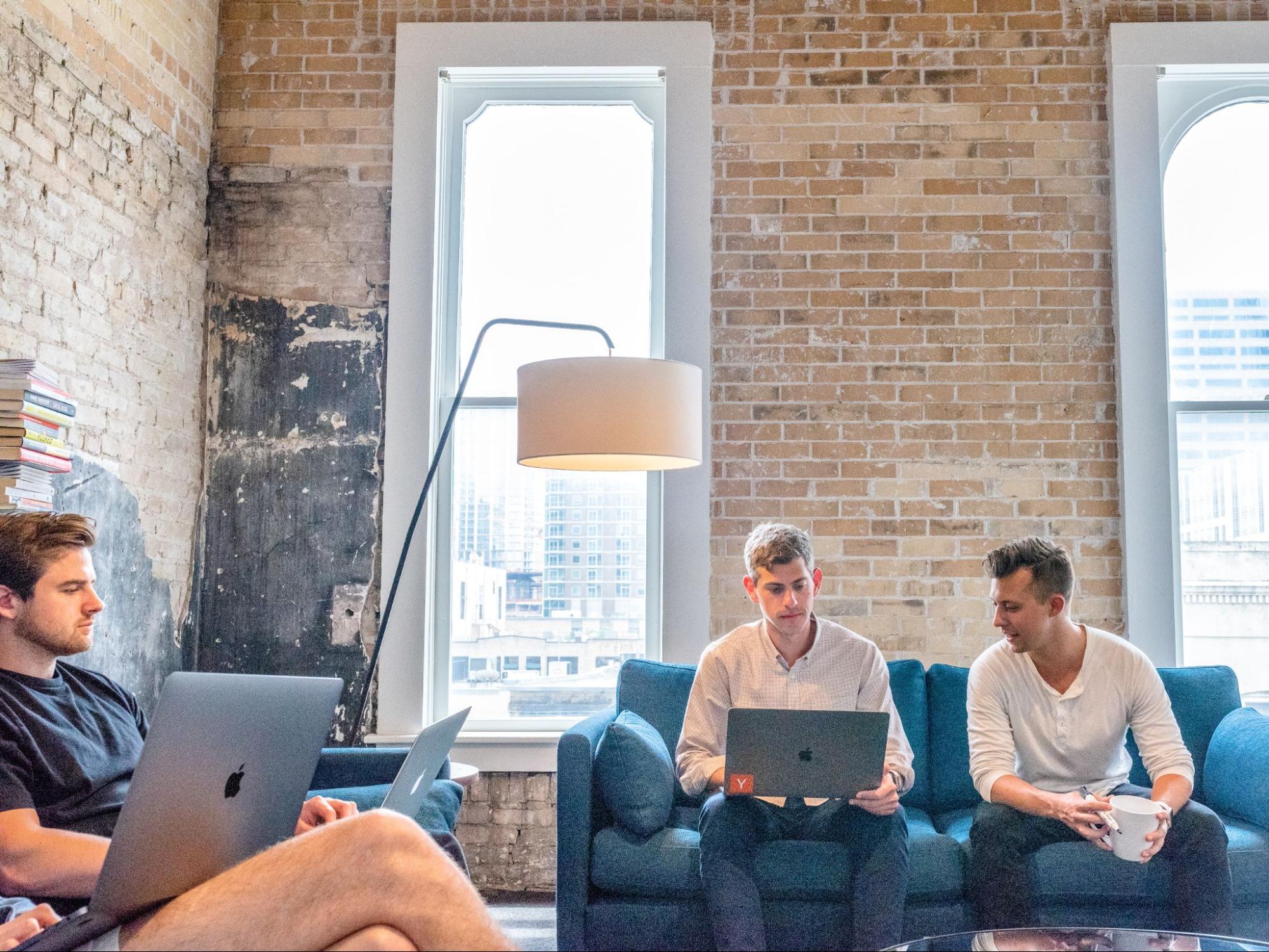 Three workers working on laptop computers, one wearing shorts and a t-shirt, while the other two have pants and long-sleeved shirts