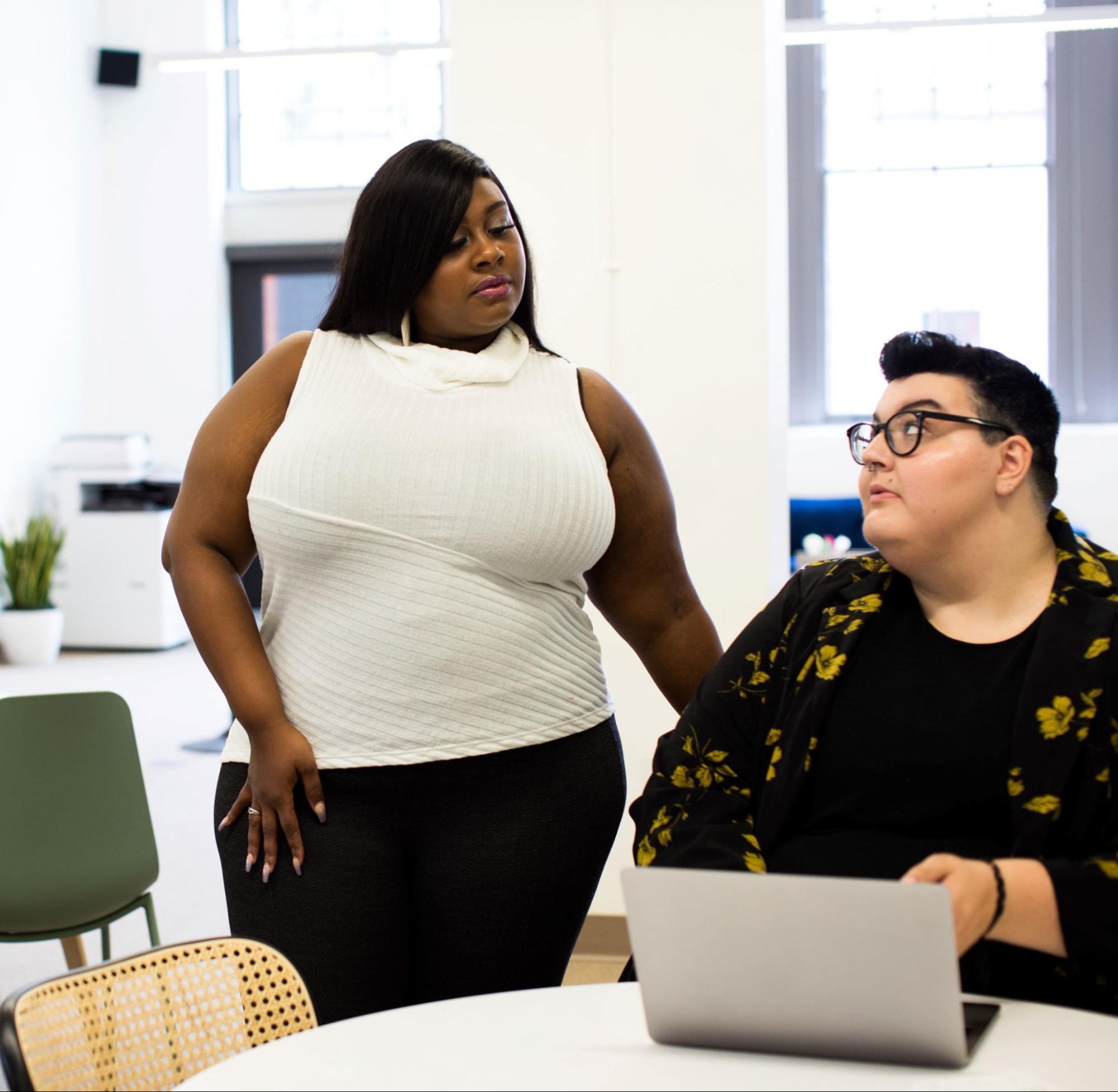 A worker sitting at their desk with a computer, looking up at another worker standing over their desk