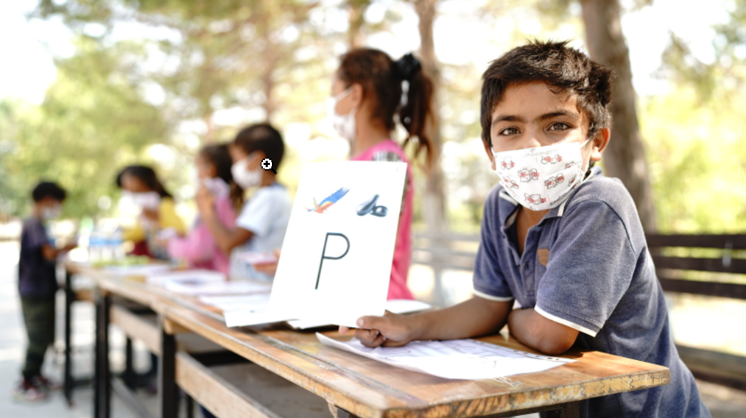 A row of desks is set-up outside under the shade of trees and elementary-age children are sitting or standing near them. There is a close-up of a young boy sitting at a desk and facing the camera, wearing a cloth mask and holding up a piece of paper with the letter "P."