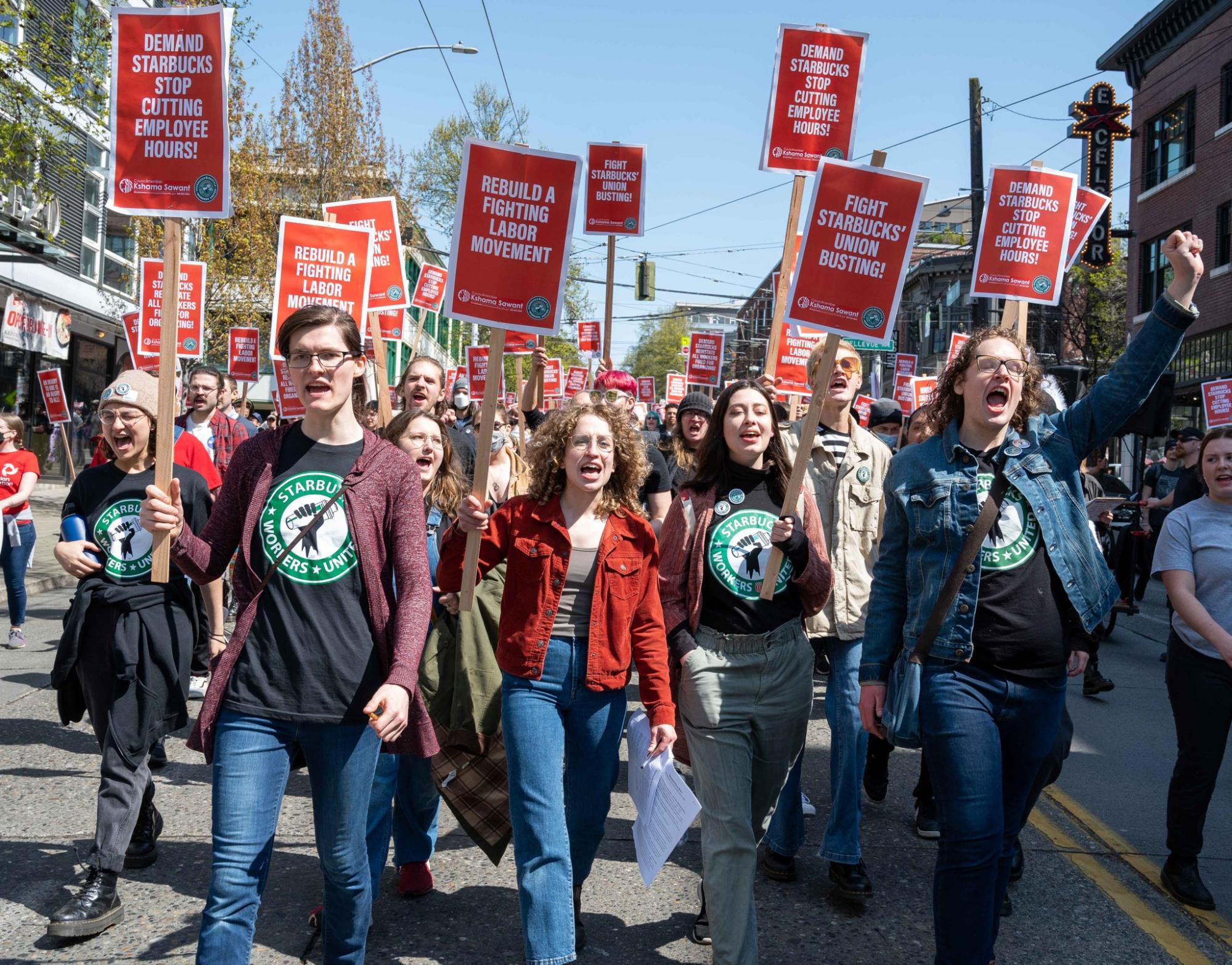 A large group of protesters walk down the middle of a street carrying signs that read, " Fight Starbucks' Union Busting!' and others.