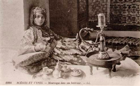A woman wearing long scarves and skirts, lounges against a wall with her legs stretched before her, feet crossed near a hookah on a table.