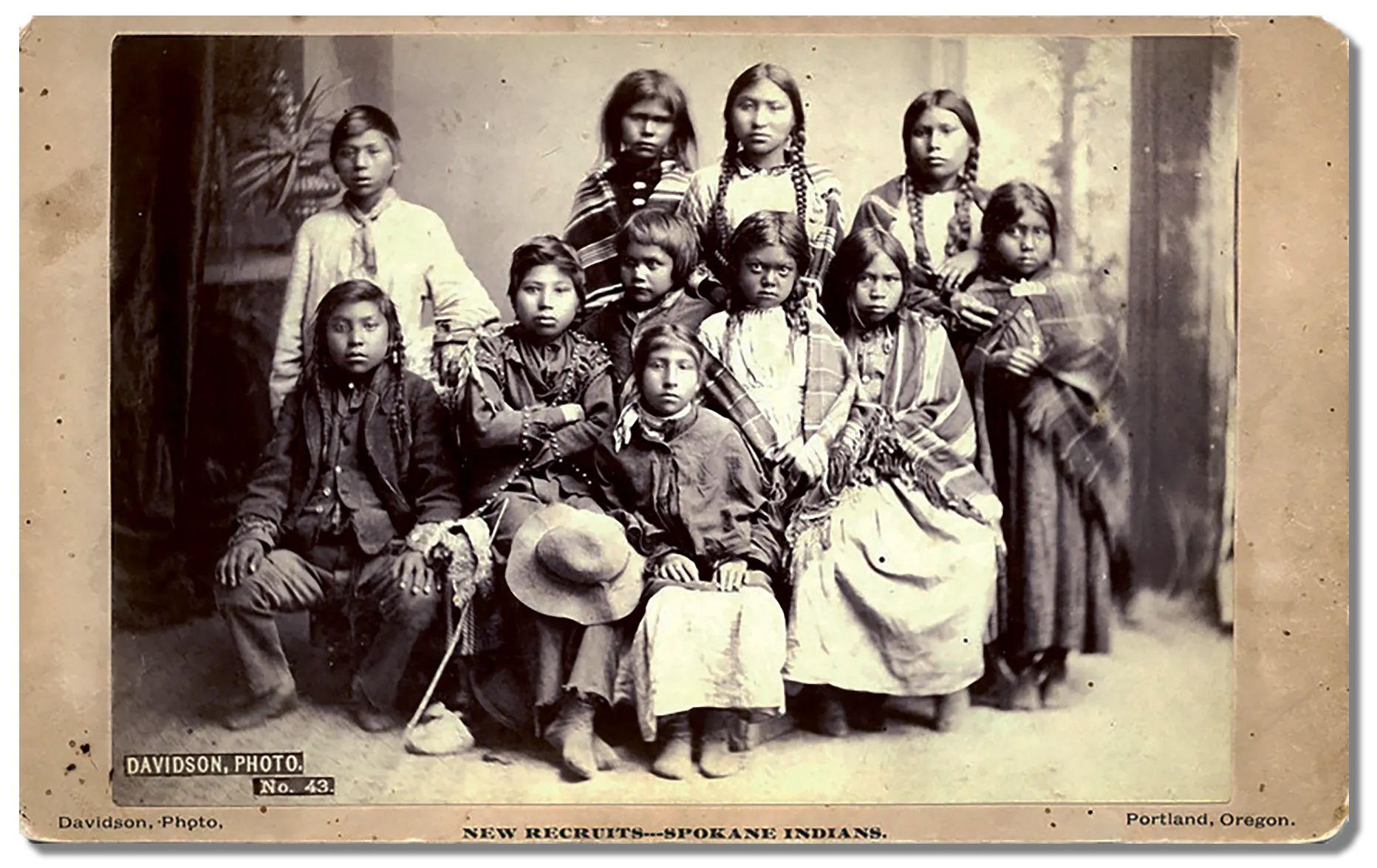 Eleven young students from the Spokane tribe are gathered together for a photo, fear and distrust is evident in their facial expressions.