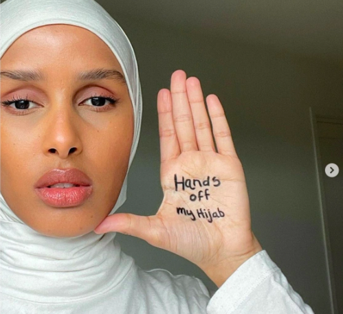 A Muslim woman wears a hijab that covers her head, neck, and arms. She holds her palm towards the camera with "Hands off my Hijab" written with black marker on it.