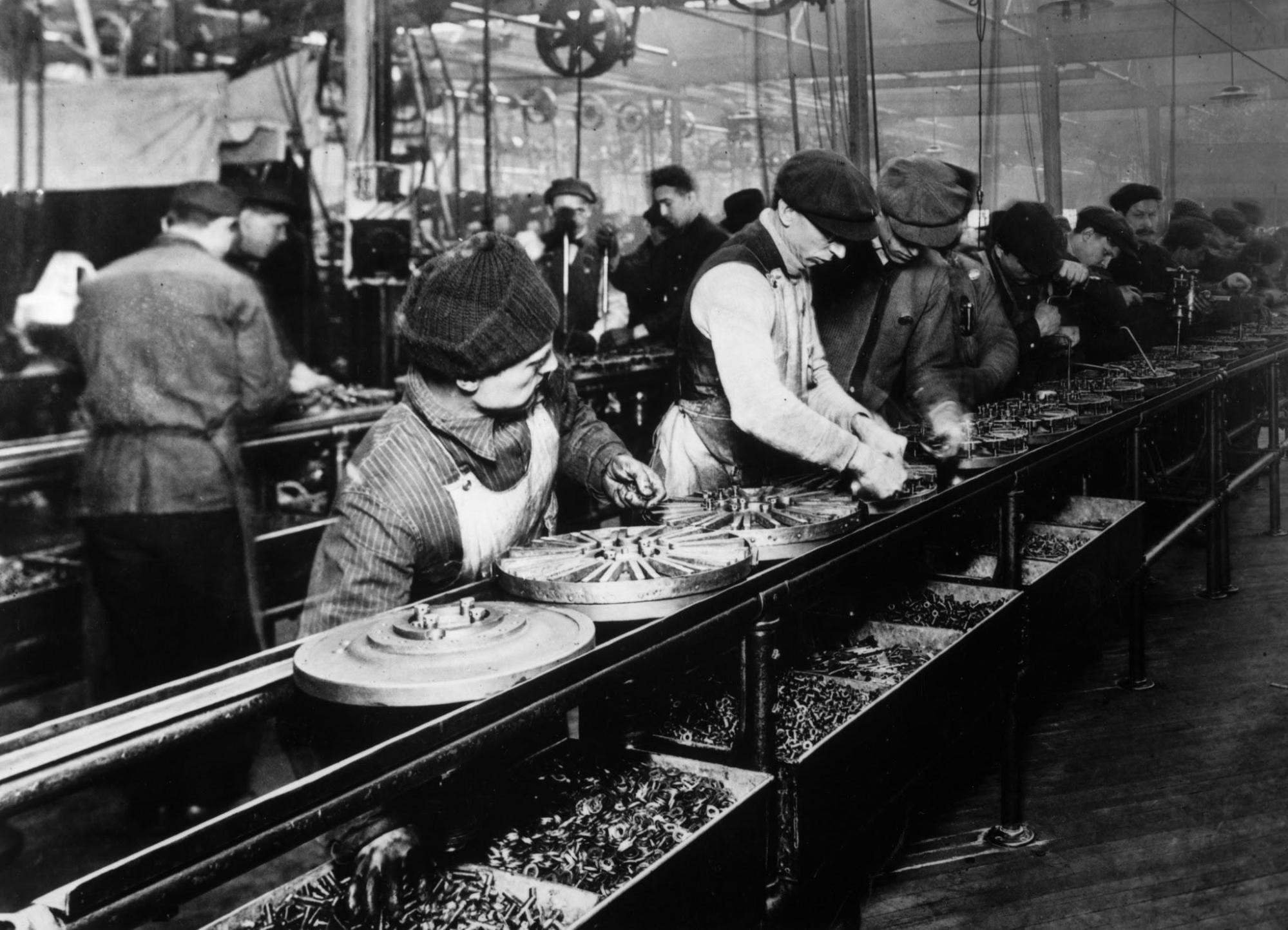 A black and white photograph shows men lined up close together working an assembly line with various parts.
