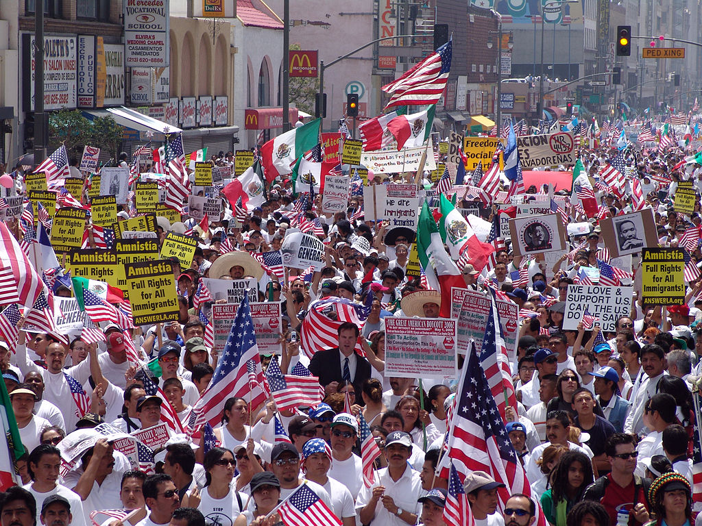 A large crowd of people fills the street, many carrying American flags and signs calling for full rights for immigrants.