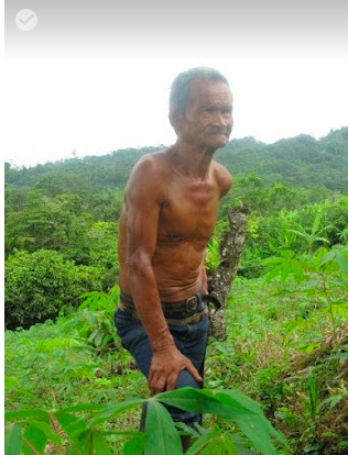 An older Columbian man is shirtless and leans his hand on his denim-covered knee. Lush greenery landscape is surrounding him.