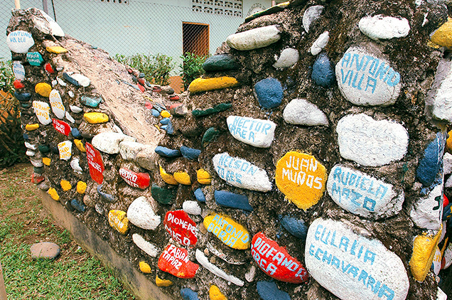 A wall is built from dirt various round stones painted red, yellow, blue, and white, with names painted on them.