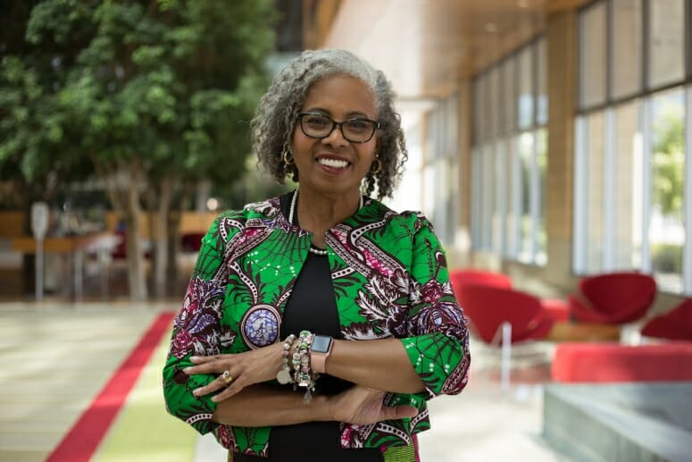 An African American woman smiles widely and faces the camera. She has black glasses, shoulder length gray hair, and wears a brightly designed jacket.