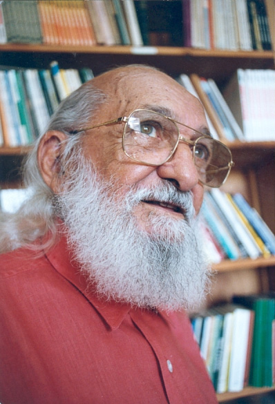 A Brazilian man looks off to the side of the camera. He has a white beard and wears glasses.