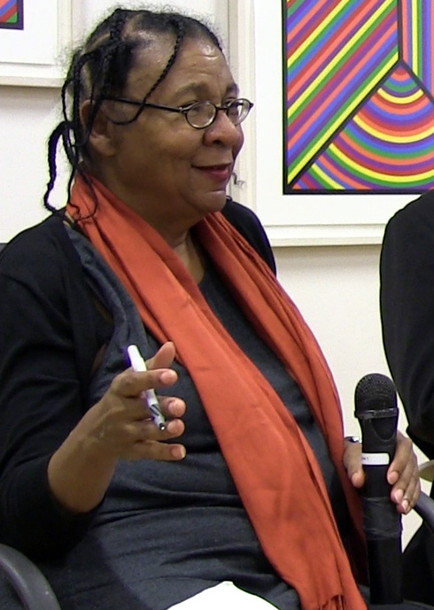 A Black woman is pictured speaking with a microphone and wearing an orange scarf around her neck.
