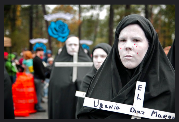 Protestors are draped in black robes and wear white makeup on their faces with red tears. They carry white crosses with names written on them.