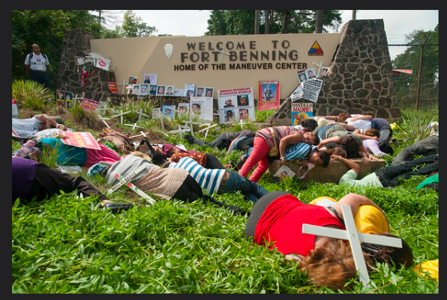 A large number of people are sprawled in the grass with white wooden crosses in front of a sign that reads, "Welcome to Fort Benning."