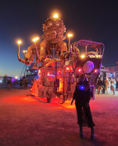 Image of person at dusk standing in front of a large art project in the desert. The art looks like a gas mask on a head with several octopus like legs emerging from the head, it is propped on a cart. There are flames emerging from the top of the head and the end of the legs