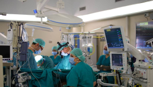six surgeons in face masks and scrubs performing surgery in an operation room. Medical equipment surrounds them.