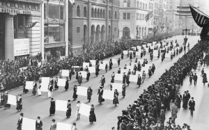 Women marching in the middle of a street in New York city in 1917 at a Women's suffragists parade. Crowds of people lined the streets. American flags flying at local businesses. Bank advertisting "be a patriot by getting bonds"