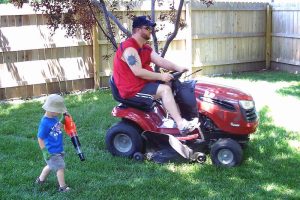 image of a man in a fenced in backyard on a riding lawn mower, mowing grass while a young boy, his son walks next to him with a leaf blower.