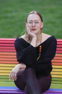 Sasha Costanza-Chock is seated on a rainbow-toned bench and smiles at the camera.