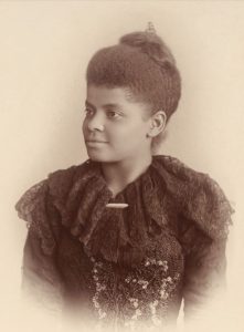 Ida B Wells is smiling slightly and wearing a dark, lacy dress with hair in a bun.