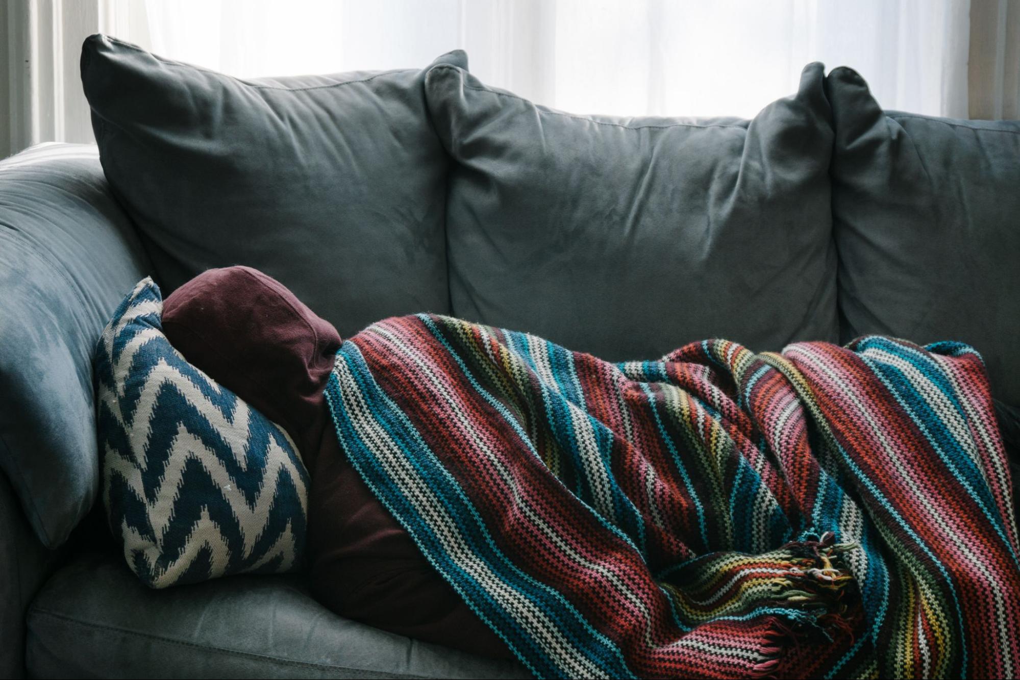 A person wearing a hooded sweatshirt is lying underneath a brightly-striped blanket on a couch.