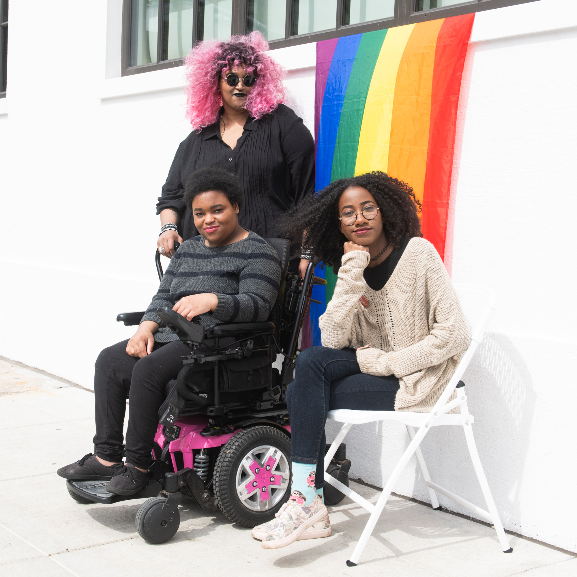 Three BIPOC people are smiling at the camera; 1 stands with a cane, another sits in a chair, and another sits in a wheelchair.