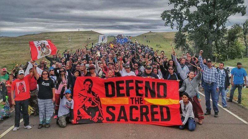 A large group of indigenous people standing behind a sign that says "Defend the Sacred"