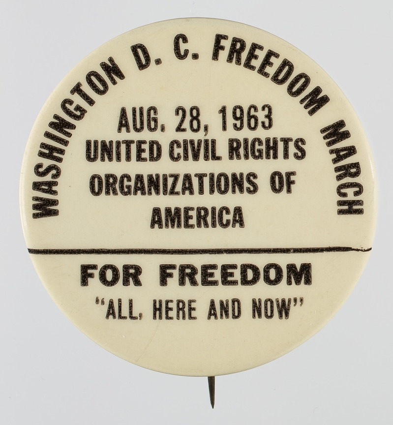 A photograph of a button from the Washington DC Freedom March reads," Aug 28, 1963, United Civil Rights Organizations of America, FOR FREEDOM, "ALL, HERE AND NOW."
