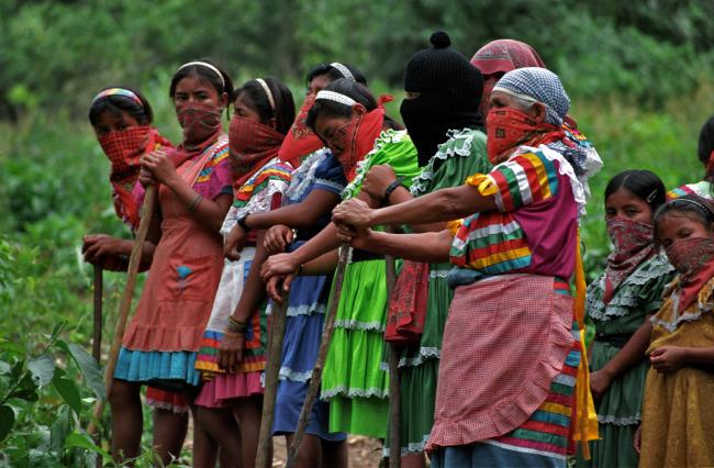 Indigenous women with their mouths covered with bandanas wearing colorful skirts and holding staffs